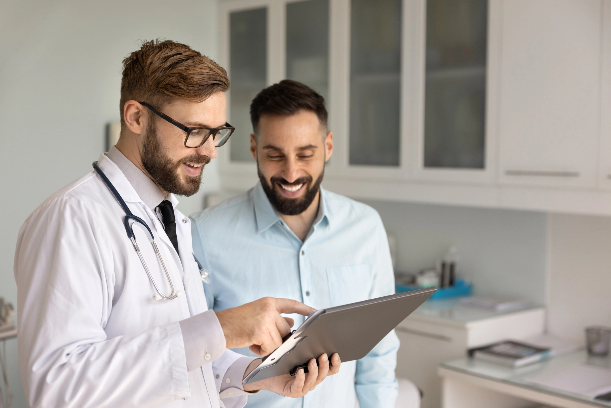 Two men in a doctor's office, both wearing glasses and stethoscopes, looking at a tablet computer