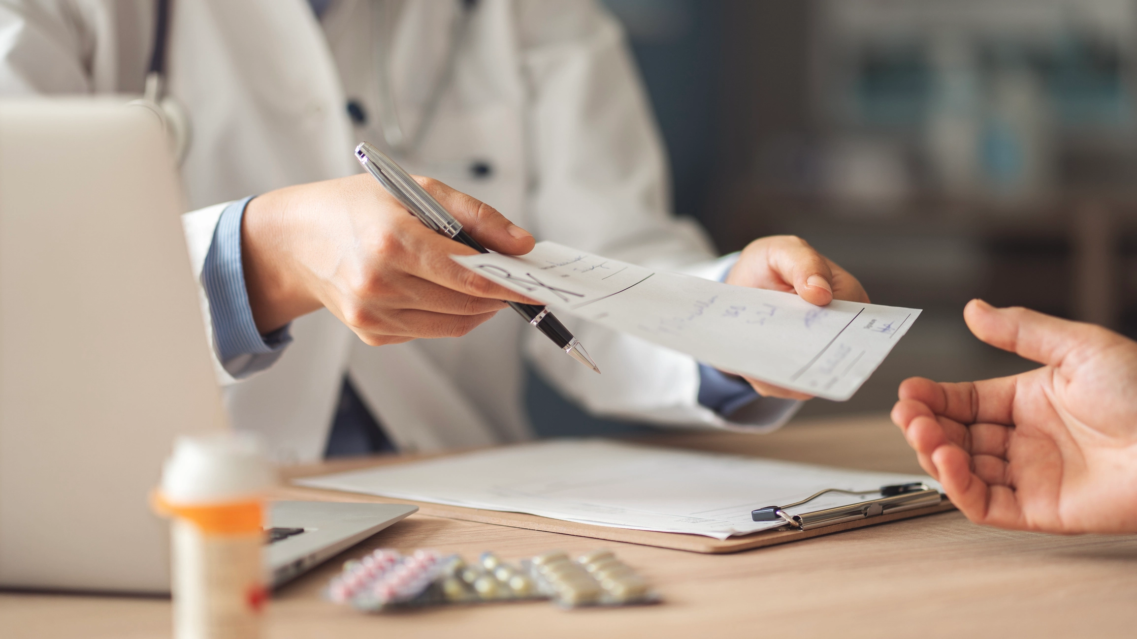 Doctor sitting at a desk with a laptop, papers, and a bottle of pills in front of them